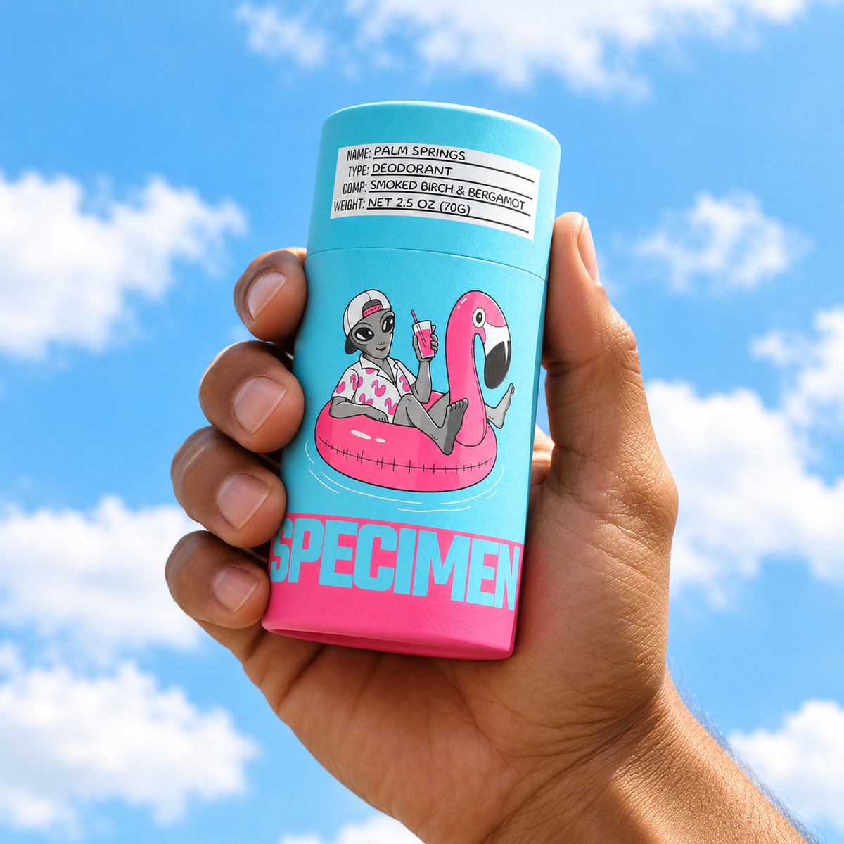 Hand holding Specimen Palm Springs aluminum-free deodorant with flamingo float alien artwork against a blue sky
